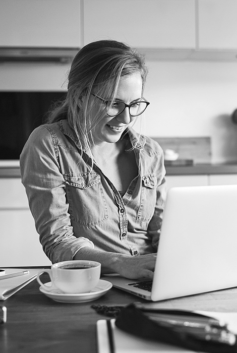 Girl with glasses sitting in her kitchen on her laptop with coffee.